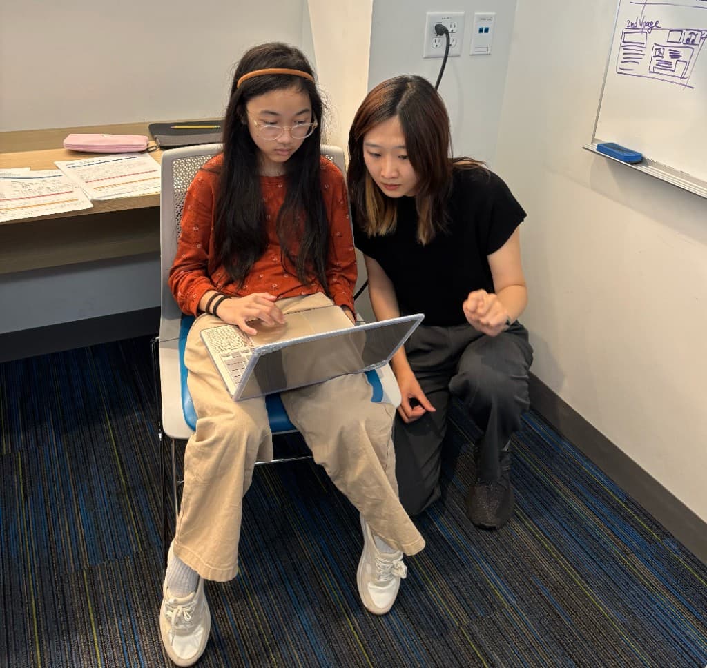 Mentor kneeling beside a student as they work together on a laptop during Game Design