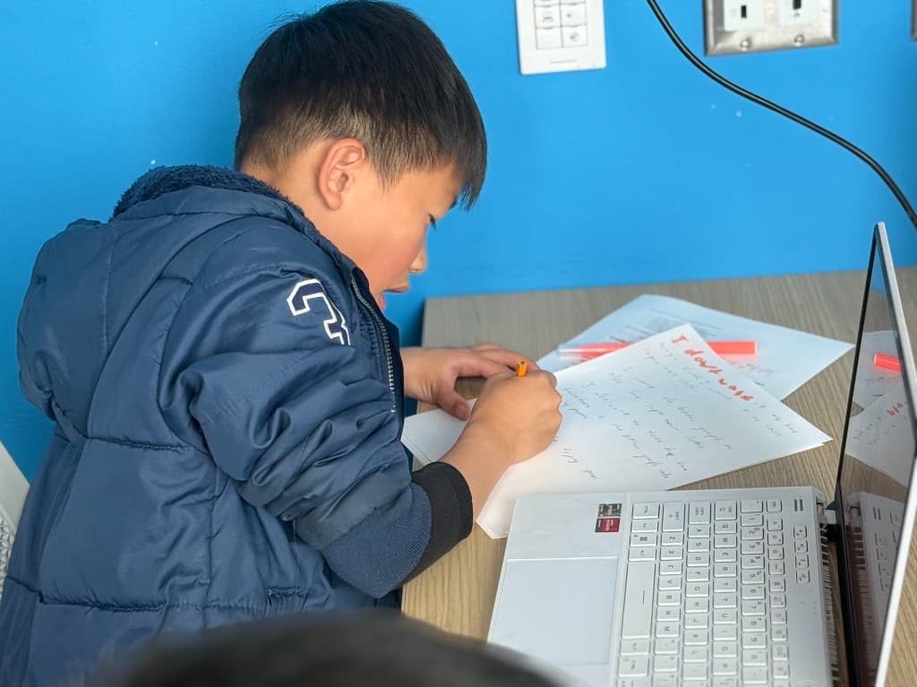 Student writing story notes at a desk next to a laptop
